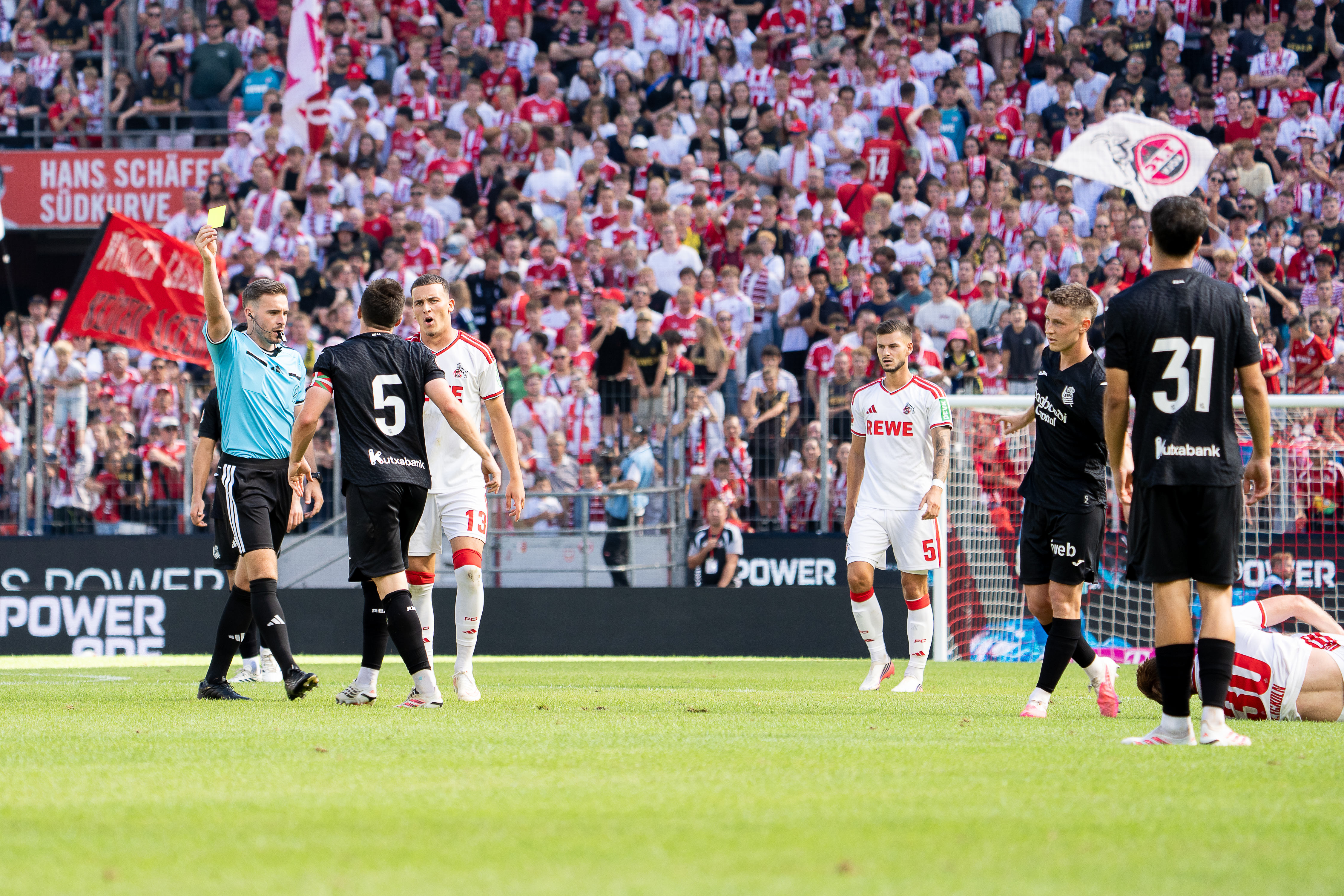 Schiedsrichter Tom Bauer zeigt Igor Zubeldia (#5; Real Sociedad) wegen Foulspiel die Gelbe Karte, umgeben von Spielern beider Teams im Freundschaftsspiel 1. FC Köln – Real Sociedad.
Foto: Alexander Franz