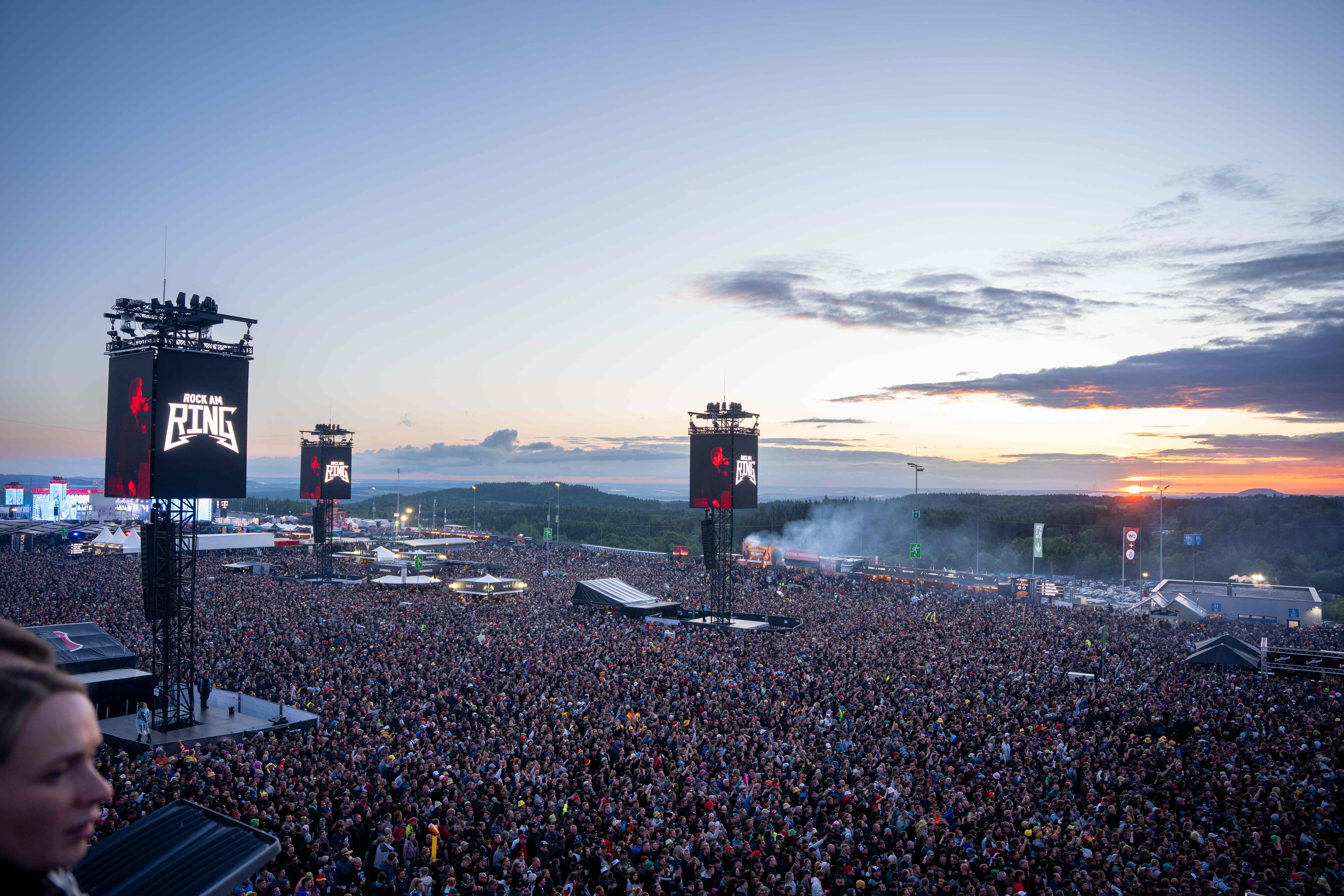 Weitwinkelaufnahme mit großer Menschenmenge beim Rock am Ring Festival am Freitag, Nürburgring. Die Bühne mit der Beschriftung Rock am Ring ist im Hintergrund sichtbar, dahinter der Sonnenuntergang am Abendhimmel.