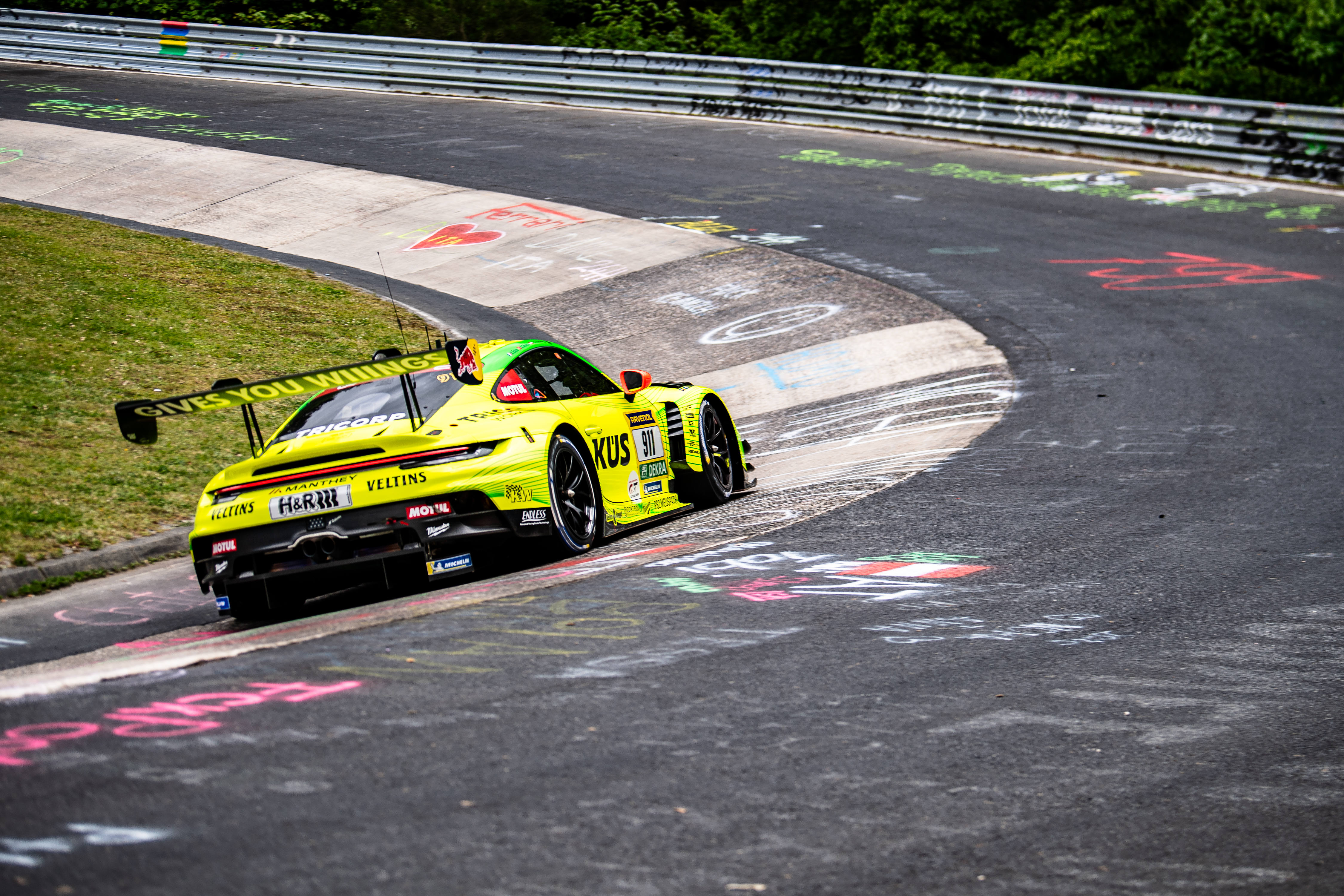 Manthey (#911) SP 9. Ein gelb-grüner Porsche 911 GT3 R mit Werbeaufschriften fährt eine Kurve der Nürburgring-Nordschleife entlang.